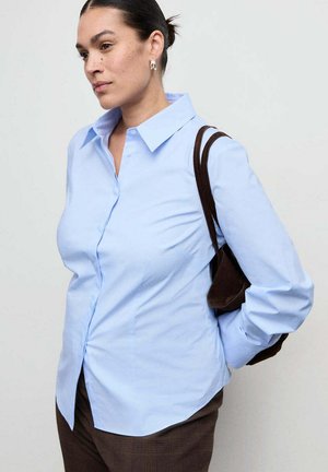 Femme aux cheveux noirs attachés en chignon porte une chemise bleu clair à boutons et un pantalon marron, portant un petit sac à bandoulière marron.