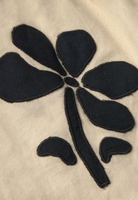 Zwarte vilt bloemenapplicatie op een beige stoffen achtergrond; drie afgeronde bloemblaadjes en een centrale stam; gestikte randen zichtbaar, wat textuur toevoegt.