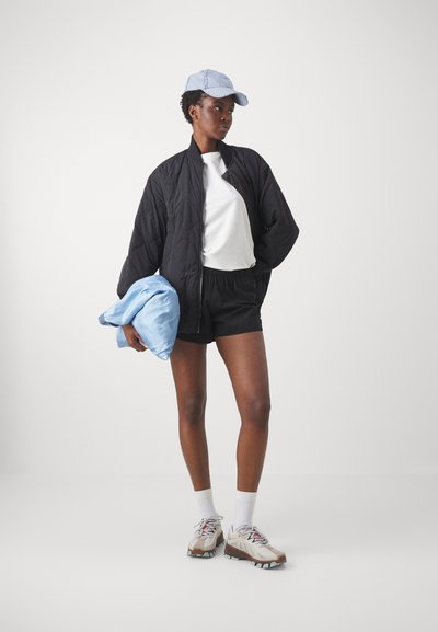 Svart quilted bomberjakke, hvit t-skjorte, svarte shorts, lyseblå caps, holder en lyseblå jakke, iført beige sneakers med grå detaljer.