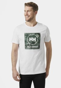 Camiseta de algodón blanca con un estampado gráfico de camuflaje verde, con el texto y el logotipo de "Helly Hansen" exhibidos de forma prominente en el centro.