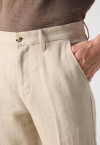 Pantalones de lino beige con corte relajado, con bolsillos laterales y cierre de un solo botón. Tejido texturizado con costuras visibles.