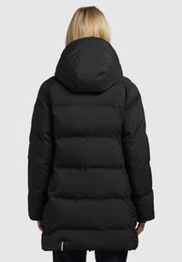 Persoon met blond haar in een lange zwarte pufferjas met een grote capuchon, gezien van achteren tegen een neutrale achtergrond.