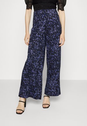 Pantalon taille haute à jambes larges avec imprimé léopard bleu et noir, associé à des sandales noires à talons et un top noir à manches en dentelle.