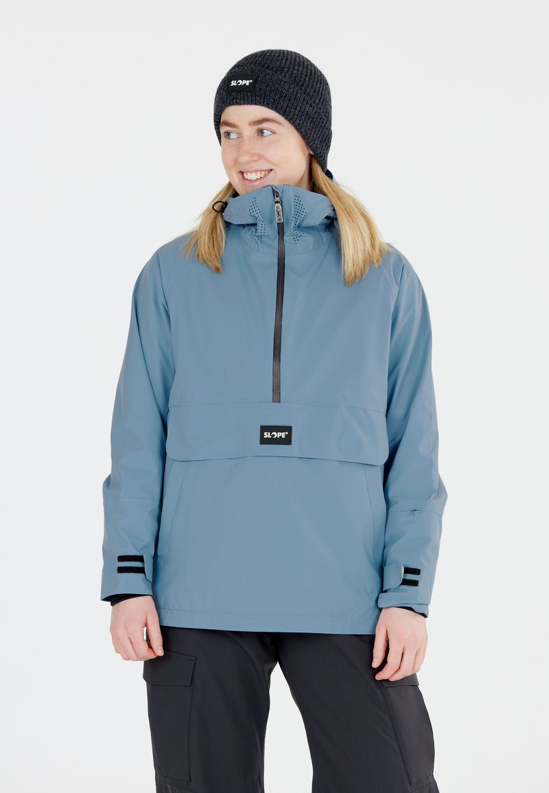 Blauer wasserdichter Pullover mit Viertelreißverschluss, Fronttasche und schwarzen Akzenten. Getragen mit einer dunklen Strickmütze. Geeignet für den Einsatz im Freien.