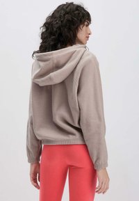 Fleece hoodie in licht taupe met een ontspannen pasvorm, trekkoord capuchon en geribbelde boorden. Gecombineerd met heldere koraalkleurige leggings.