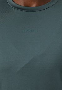 Tričko s krátkym rukávom, priliehavé, v tmavej teal farbe, vyrobené z hladkej tkaniny. Na hrudi sa nachádza vyvýšené logo "BORG".