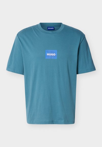 Къса тениска в тюркоазено синьо с кръгло деколте и центрирано синьо квадратче с логото "HUGO" на гърдите.