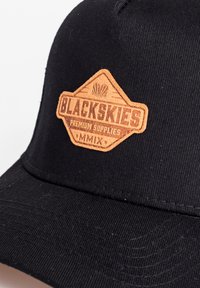 Căciulă neagră cu un patch din piele maro texturată pe care este embosat "BLACKSKIES PREMIUM SUPPLIES MMIX" într-un design hexagonal.