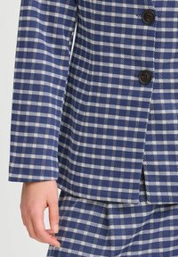 Blazer à carreaux bleus et blancs avec des boutons foncés, porté sur une jupe assortie à carreaux, la main gauche visible reposant le long du corps.