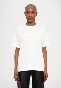 Λευκό βαμβακερό μπλουζάκι με στρογγυλή λαιμόκοψη και κοντά μανίκια, με μια διακριτική ανάγλυφη λεπτομέρεια λογότυπου στο στήθος. Φο ριέται με μαύρο δερμάτινο παντελόνι.