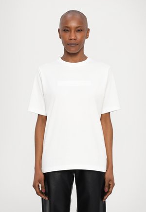 White cotton t-shirt with a round neck and short sleeves, featuring a subtle embossed logo detail across the chest. Worn with black leather pants.