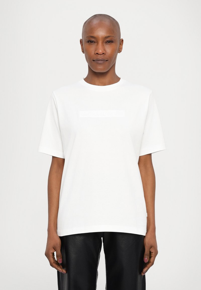 Λευκό βαμβακερό μπλουζάκι με στρογγυλή λαιμόκοψη και κοντά μανίκια, με μια διακριτική ανάγλυφη λεπτομέρεια λογότυπου στο στήθος. Φοριέται με μαύρο δερμάτινο παντελόνι.