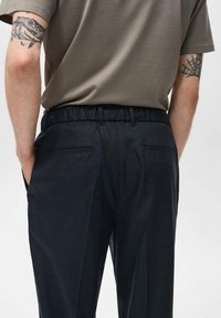 Zwarte, elastische broeken met zijzakken, een gladde textuur en een tailored pasvorm. Gedragen met een grijze, shirt met korte mouwen.