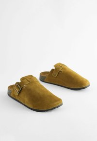 Brune ruskindsklipklapper med rund tå, der har en justerbar rem og en plettet sort sål. Glat tekstur og minimalistisk design.