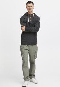 Sudadera gris oscuro con detalles naranjas y botones de madera, combinada con pantalones cargo verde claro y zapatillas blancas. Fondo neutro.