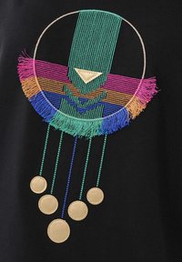 Vêtement noir brodé avec un motif circulaire en vert, rose et bleu orné de franges, agrémenté de détails circulaires dorés suspendus en dessous.