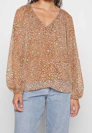 Kvinde iført en brun gennemsigtig langærmet bluse med grønne og hvide abstrakte blomsterprint og lyseblå jeans.