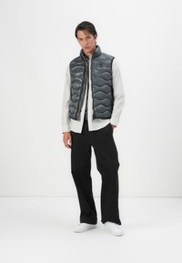 Gray quilted puffer vest with zipper, featuring a subtle wave pattern. Worn over a white striped shirt and paired with black pants and white shoes.