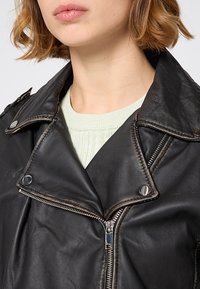 Czarna skórzana kurtka motocyklowa z srebrnymi zamkami i zatrzaskami, z szerokim kołnierzem i teksturowanym wykończeniem. Nosi ją na jasnoszarym swetrze.