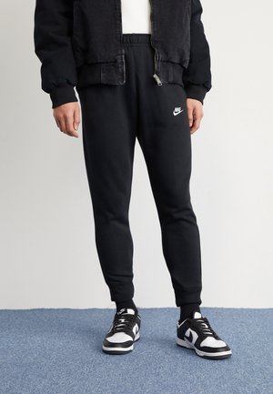 Personne portant un pantalon de survêtement Nike noir, des baskets Nike noires et blanches, une veste noire et des chaussettes noires, debout sur un tapis bleu.