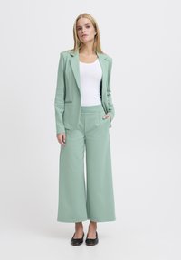 Muntgroene blazer en wijde broek van glad, zacht materiaal. De blazer heeft een enkele pocket en wordt gedragen over een witte tanktop en zwarte ballerina's.