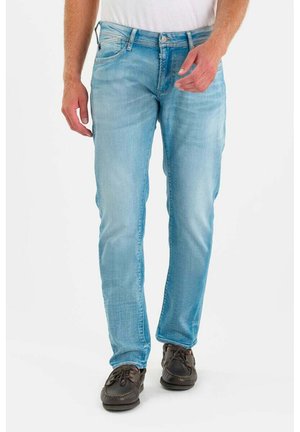 Homme portant un jean bleu clair et des chaussures à lacets marron foncé, debout devant un fond blanc uni.