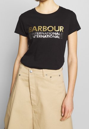Kobieta ubrana w czarny t-shirt Barbour International z złotym i srebrnym napisem, w połączeniu z beżowymi spodniami z wysokim stanem.