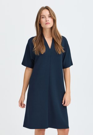 Vrouw met lang bruin haar, gekleed in een loszittende donkerblauwe jurk tot op de knie met korte mouwen en een V-hals, die voor een witte achtergrond staat.