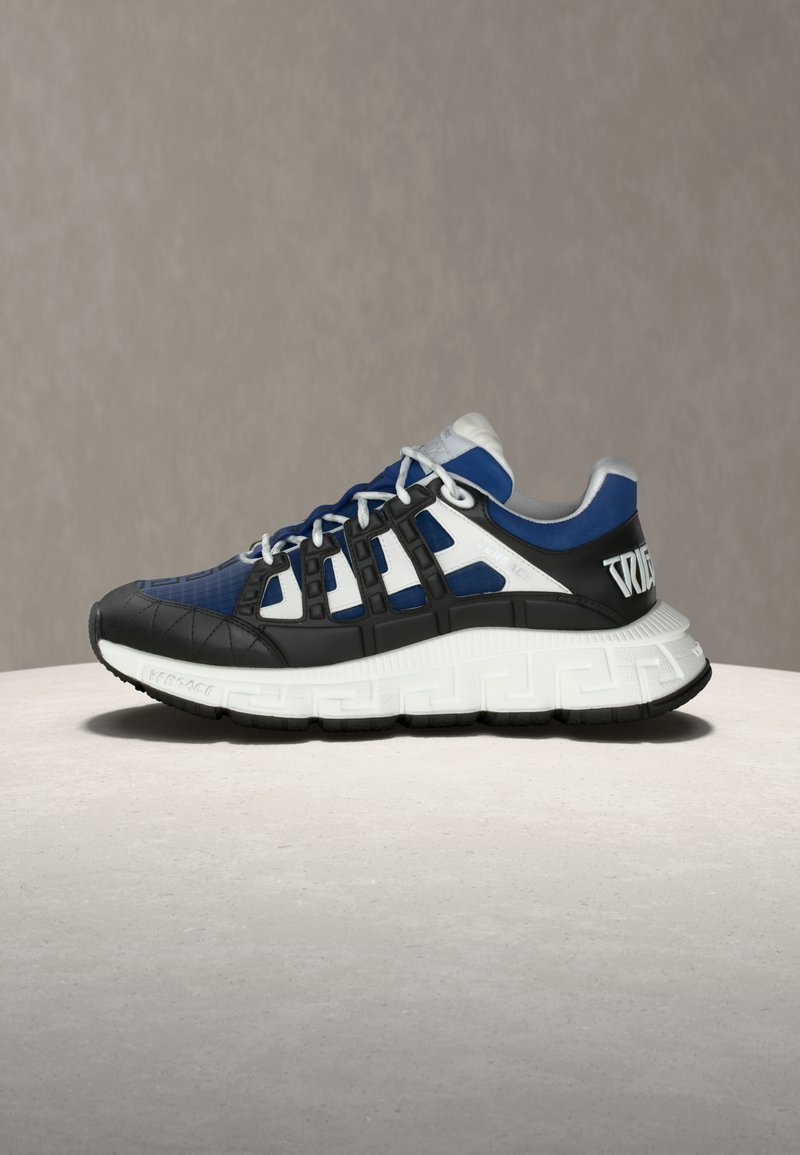 Baskets athlétiques bleu, noir et blanc avec une semelle blanche épaisse ornée de motifs géométriques, un design en cage noire et des lacets blancs sur une surface neutre.