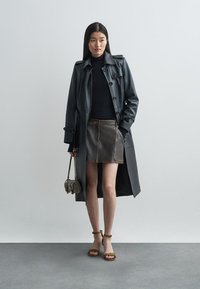 Abrigo de tren negro de cuero con botones, usado sobre una blusa negra ajustada y una falda de mezclilla marrón. Combinado con sandalias marrones y un bolso pequeño.