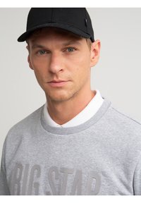 Ung mand med lys hud og grønne øjne iført en sort baseballkasket, grå sweatshirt med teksten "BIG STAR" og hvid skjorte med krave under.