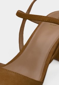 Καφέ σουέντ σανδάλια με ψηλό τακούνι, με ένα λεπτό λουράκι πάνω από τα δάχτυλα και ένα ρυθμιζόμενο λουρί στον αστράγαλο, με λείο, γυαλιστερό εσωτερικό πάτο.