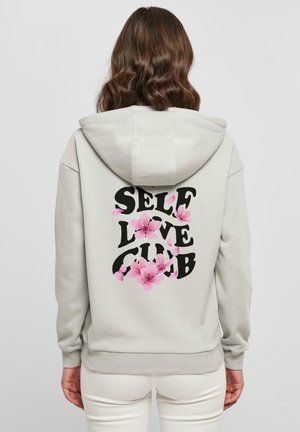 Kvinde iført en lysegrå hættetrøje med teksten "SELF LOVE CLUB" og lyserøde blomster på ryggen, stående mod en ensfarvet baggrund.