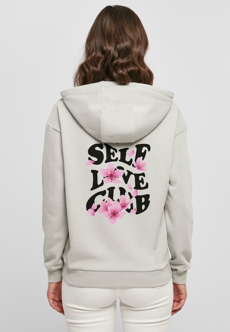 Femme portant un sweat à capuche gris clair avec le texte "SELF LOVE CLUB" et des graphiques de fleurs roses au dos, debout devant un fond uni.