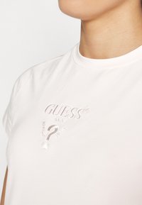 Lys rosa bomull t-skjorte med korte ermer som har innpreget "GUESS" logo og spørsmåls-tegn i en litt mørkere nyanse.