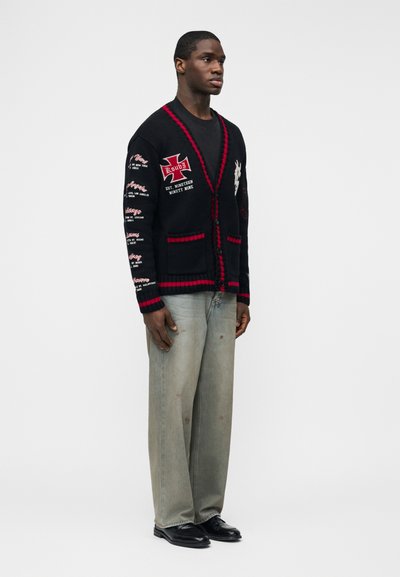 Uomo che indossa un cardigan nero con bordo rosso e toppe di testo bianche, maglia nera, jeans sbiaditi e larghi, e scarpe eleganti nere, in piedi di lato.