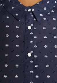 Chemise bleu marine avec boutons blancs à quatre trous et petits motifs d'abeilles blanches brodés sur tout le tissu.