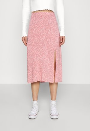 Femme portant une jupe midi rose à petits motifs floraux blancs avec une fente latérale, associée à des baskets blanches montantes et un haut blanc à manches longues raccourci.