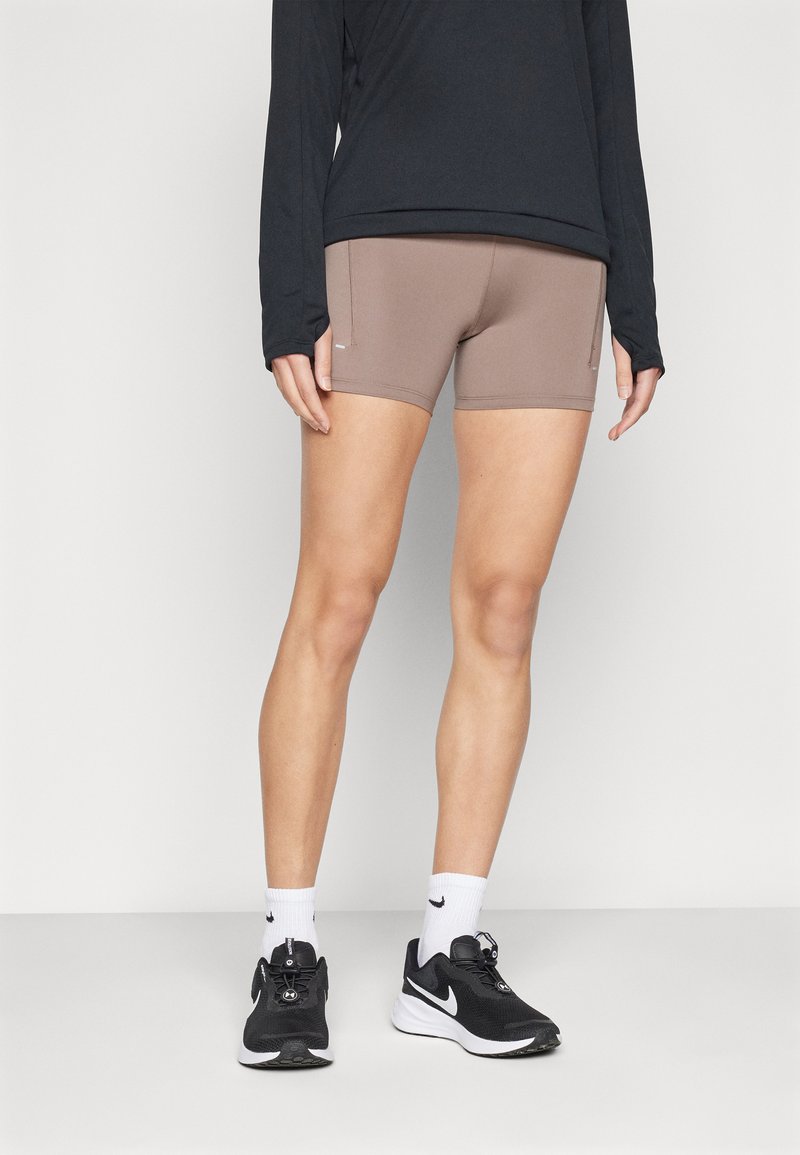 Pantalones cortos atléticos beige con un diseño ajustado, bolsillos laterales, combinados con una camiseta negra de manga larga y zapatillas de correr negras, con calcetines blancos.
