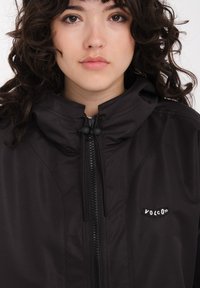 Veste à capuche noire avec fermeture éclair sur le devant, fabriquée en matériau lisse. Comprend un patch logo "Volcom" sur la poitrine et des cordons élastiques au col.