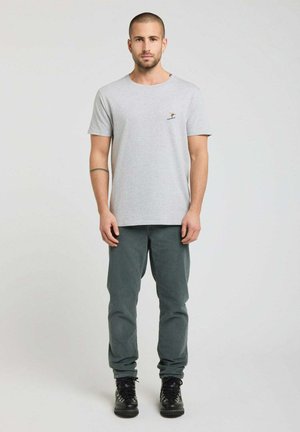 Homme debout face à l’avant, portant un t-shirt gris clair avec un petit logo de skieur, pantalon gris et bottes noires sur fond uni.
