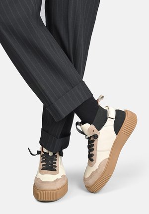 Ben iført sorte bukser med striber og sorte sokker med beige og hvide sneakers med brune tykke gummisåler.