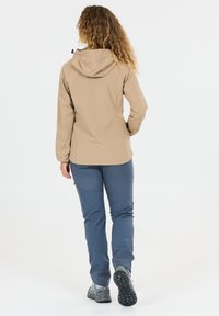 Frau mit lockigem Haar, die eine beige Kapuzenjacke, blaue Wanderhose und graue Wanderschuhe trägt, von hinten gesehen, beim Gehen.