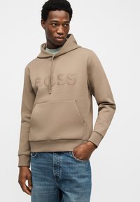 Νέος άνδρας που φοράει μπεζ φούτερ με ανάγλυφη επιγραφή "BOSS" και μπλε τζιν, στέκεται με το ένα χέρι στην τσέπη, μπροστά σε απλό φόντο.