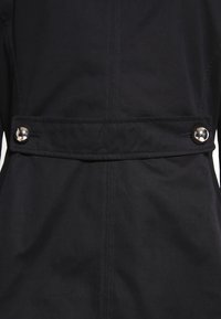 Svart kappa med en mjuk textur, insvängd midja med bälte och silverknappdetaljer. Ren linjeföring med minimalistiska designinslag.