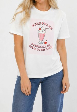 Femeie purtând un tricou alb cu grafică roz de milkshake și textul „Milkshake brings all the girls to the yard”, asortat cu blugi albaștri.