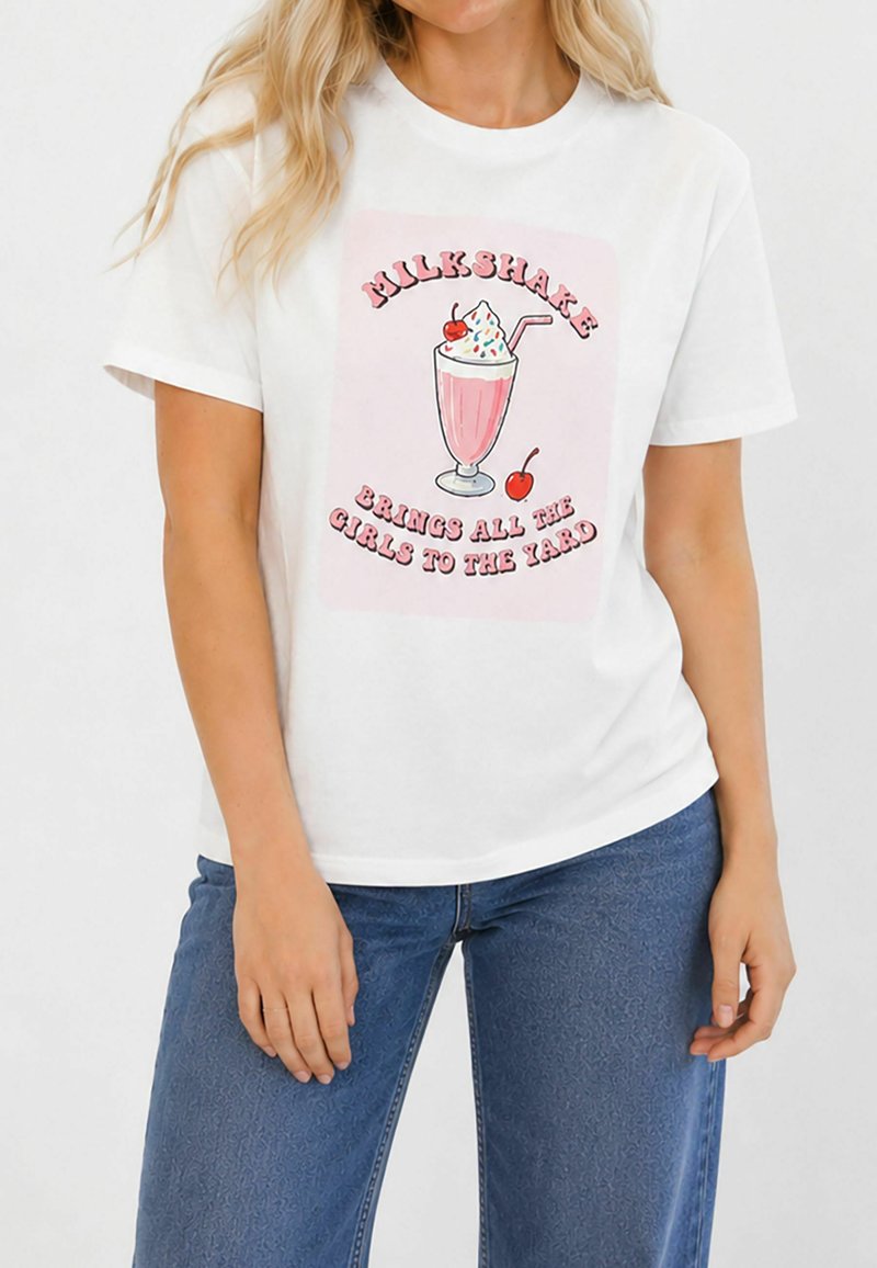 Γυναίκα με λευκό μπλουζάκι με ροζ εικόνα milkshake και κείμενο «Milkshake brings all the girls to the yard», σε συνδυασμό με μπλε τζιν.
