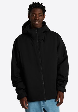 Ung mand iført en sort hættejakke med lynlås og lyseblå jeans, stående mod en ensfarvet lys baggrund.