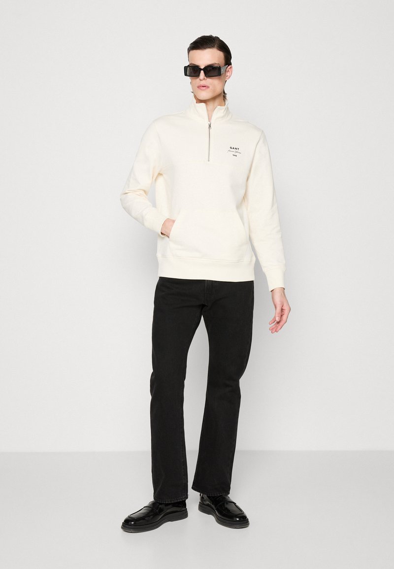 Felpa color crema con zip a quarto, tasca frontale e logo ricamato, abbinata a jeans neri e scarpe nere lucide.