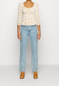 Cremetop mit Blumenmuster, quadratischem Ausschnitt, gepufften Ärmeln; kombiniert mit hellblauen, gerade geschnittenen Jeans und braunen Stiefeln mit Absatz.